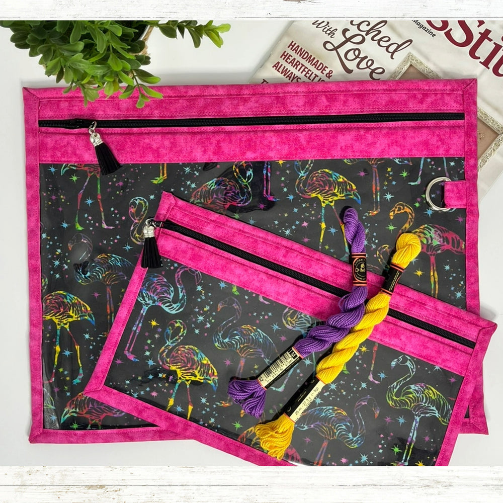 Two pink bags with black zippers on a dark fabric background with colorful flamingo pattern, accompanied by yarn.