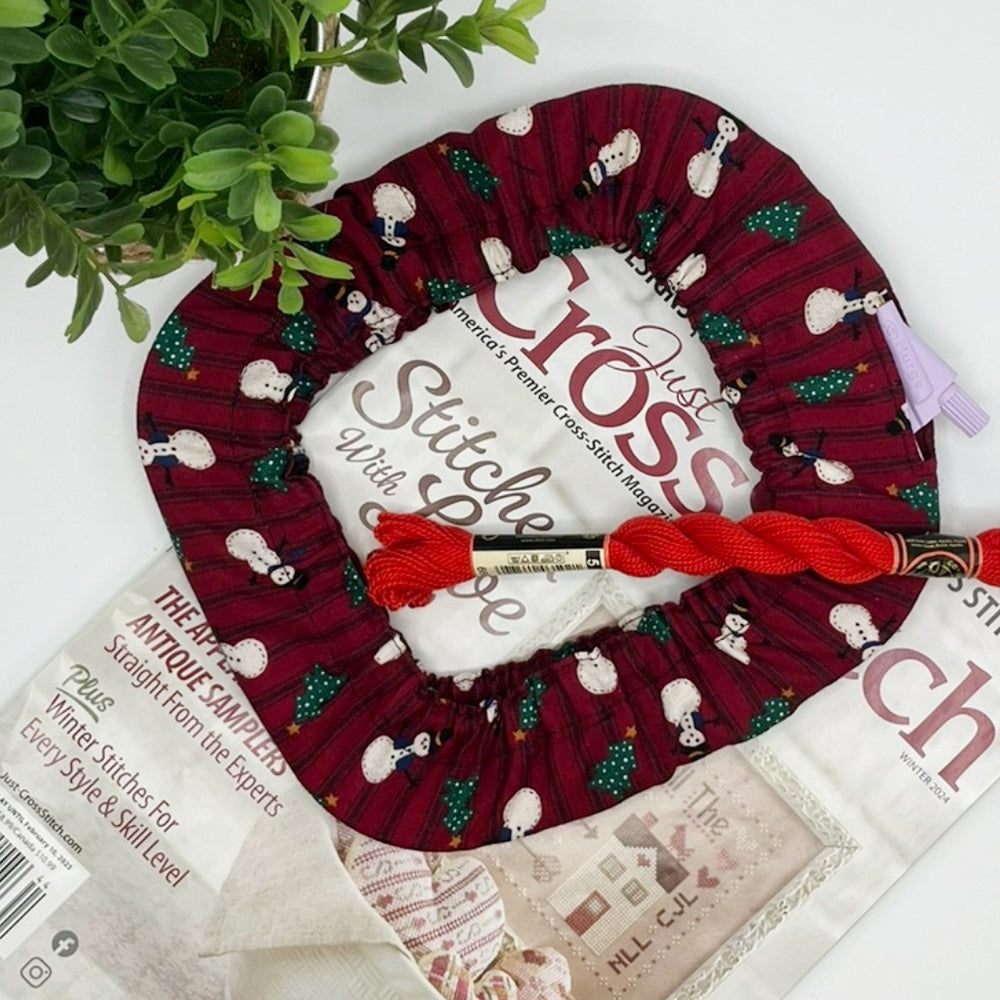 Cross stitch hoop grime guard cover in snowman theme fabric on red background with christmas trees.