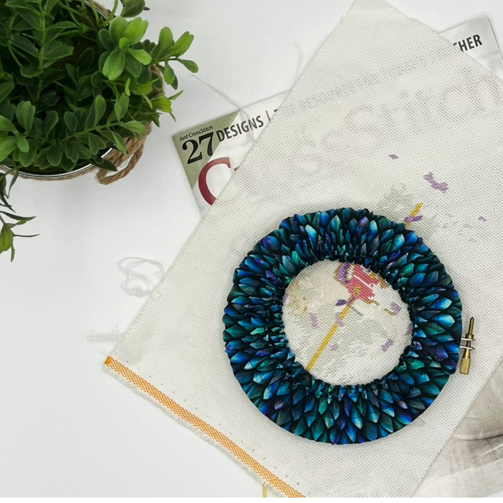 Embroidery hoop with blue and green fabric on a white surface with a plant and magazine.